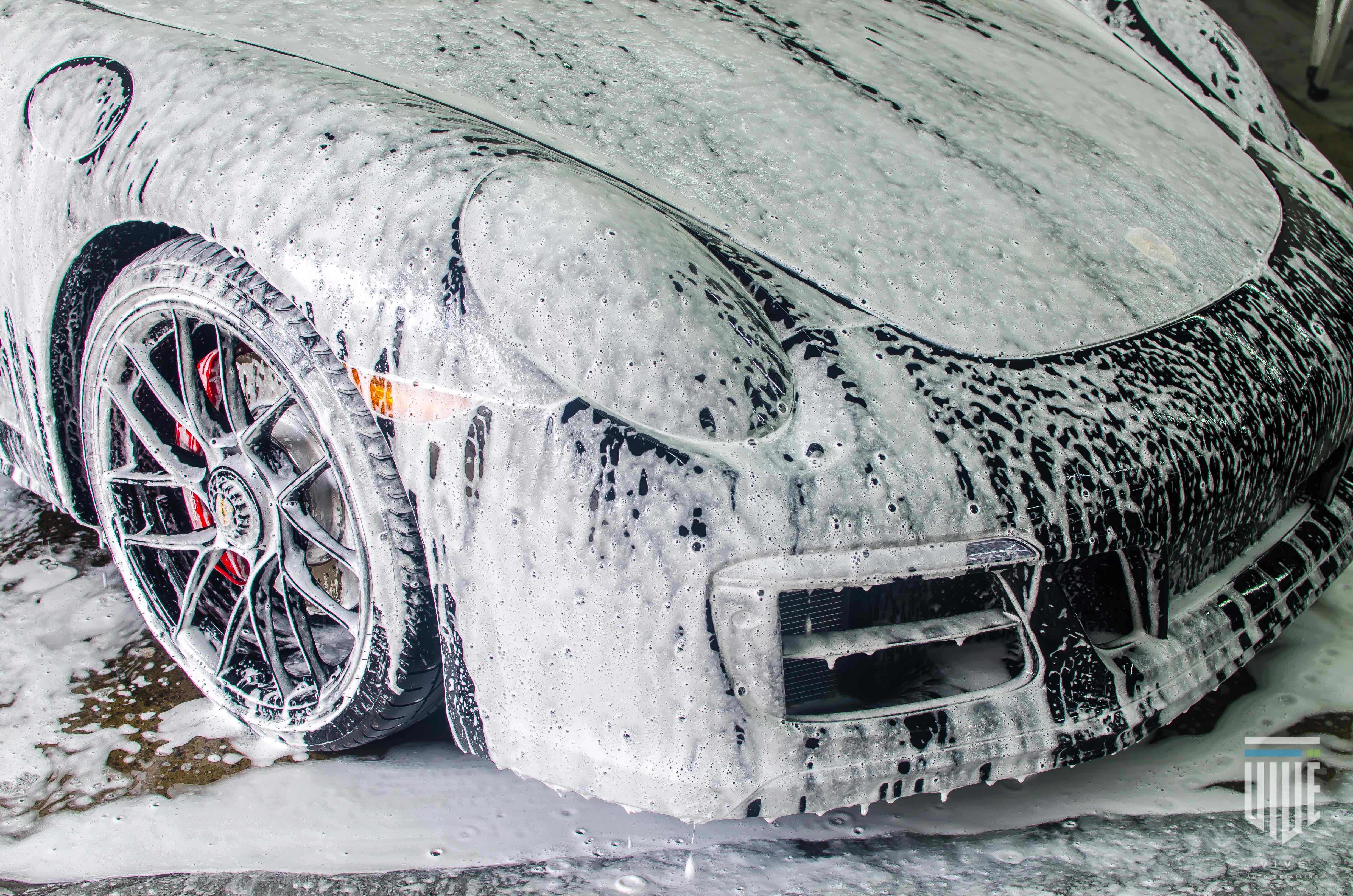 Thick white cleaning foam covers the front of a black sports car during a wash.