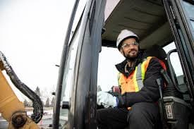 Smiling construction worker in safety gear sitting inside the cab of a yellow excavator.