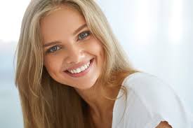 Radiant young woman with long blonde hair smiling broadly while wearing a simple white top.